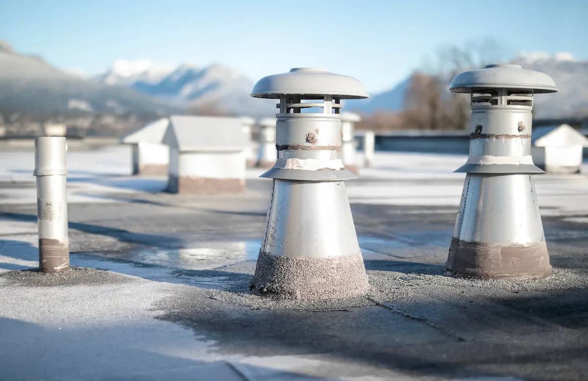 Frost-covered rooftop with several metal chimneys and vents. Snow-capped mountains and blurred trees are in the background under a clear blue sky.