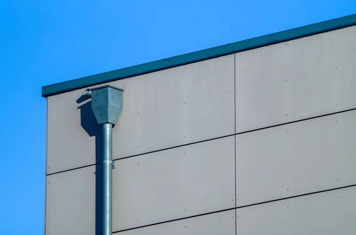 A minimalist image shows the corner of a modern building with a smooth beige facade and a steel drainpipe. The clear blue sky provides a contrasting background, highlighting the building's geometric design.