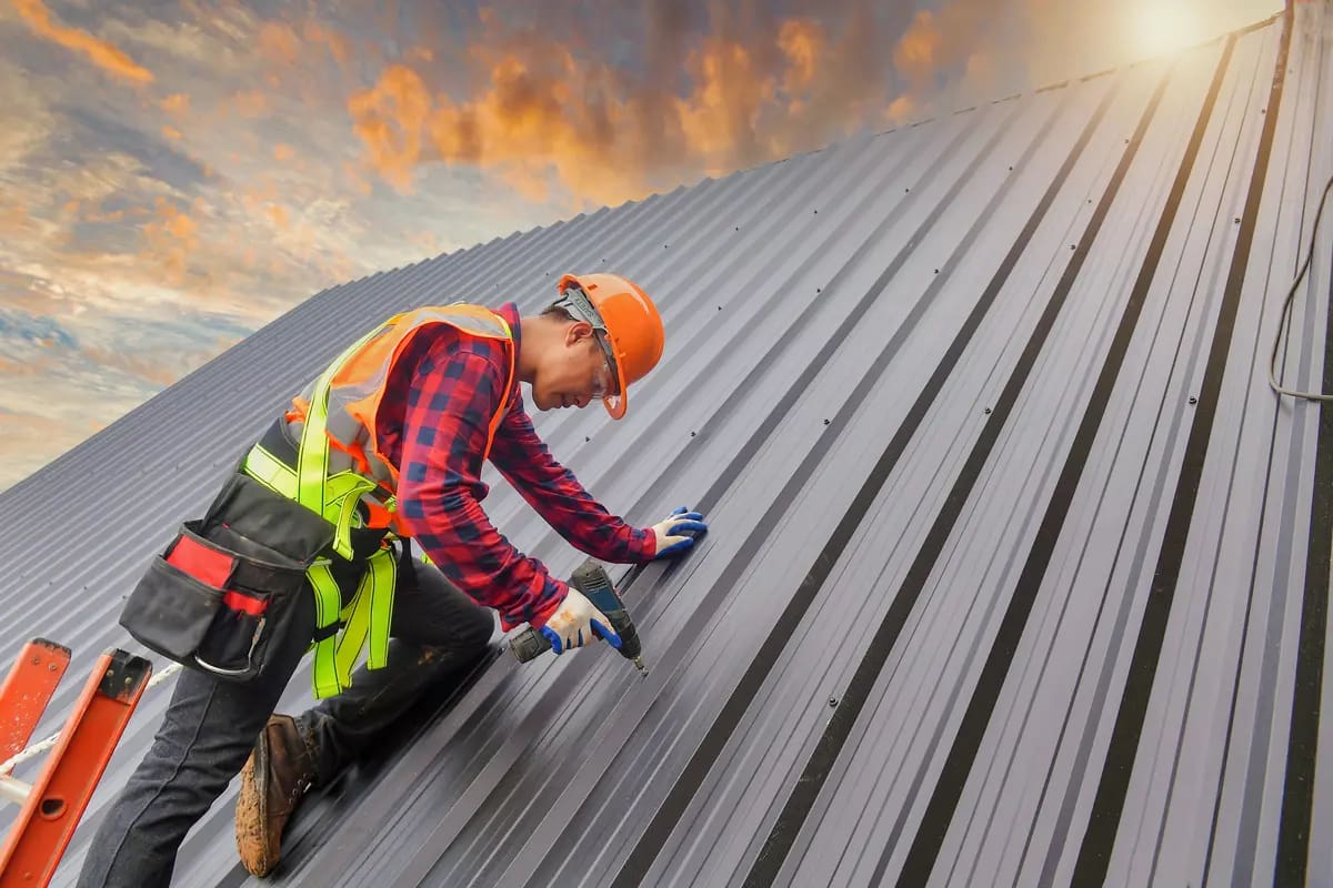 A construction worker in a safety vest and helmet kneels on a metal roof, using a tool to secure it. The sky is partly cloudy with a warm glow from the setting sun. A ladder is visible in the foreground.