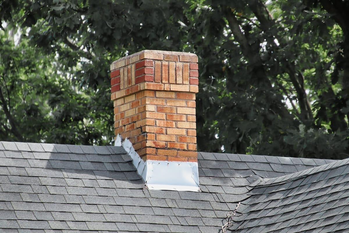 A brick chimney with varied shades of red and beige bricks extends from a shingled roof. The roof is dark gray, and there are trees with green leaves in the background. A metal flashing surrounds the base of the chimney.