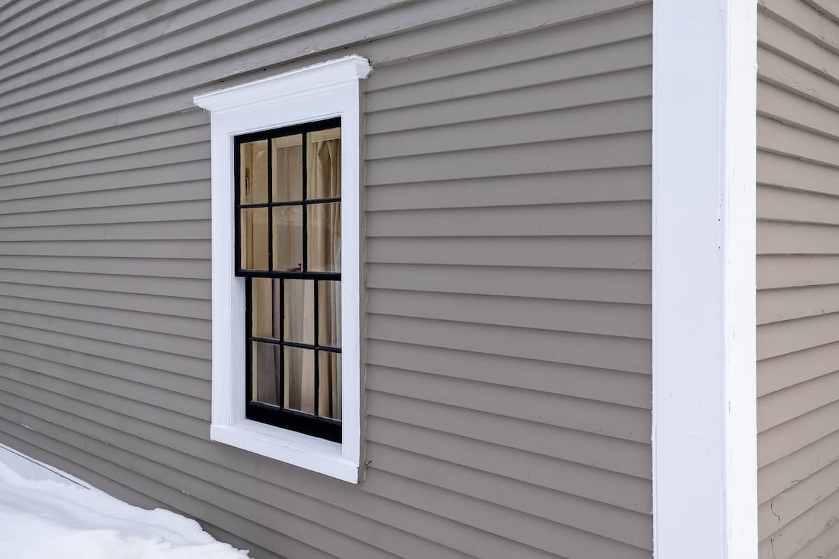A black-framed window with white trim is set in a gray, horizontally paneled wall. Snow covers the ground below, providing a stark contrast to the building's exterior.