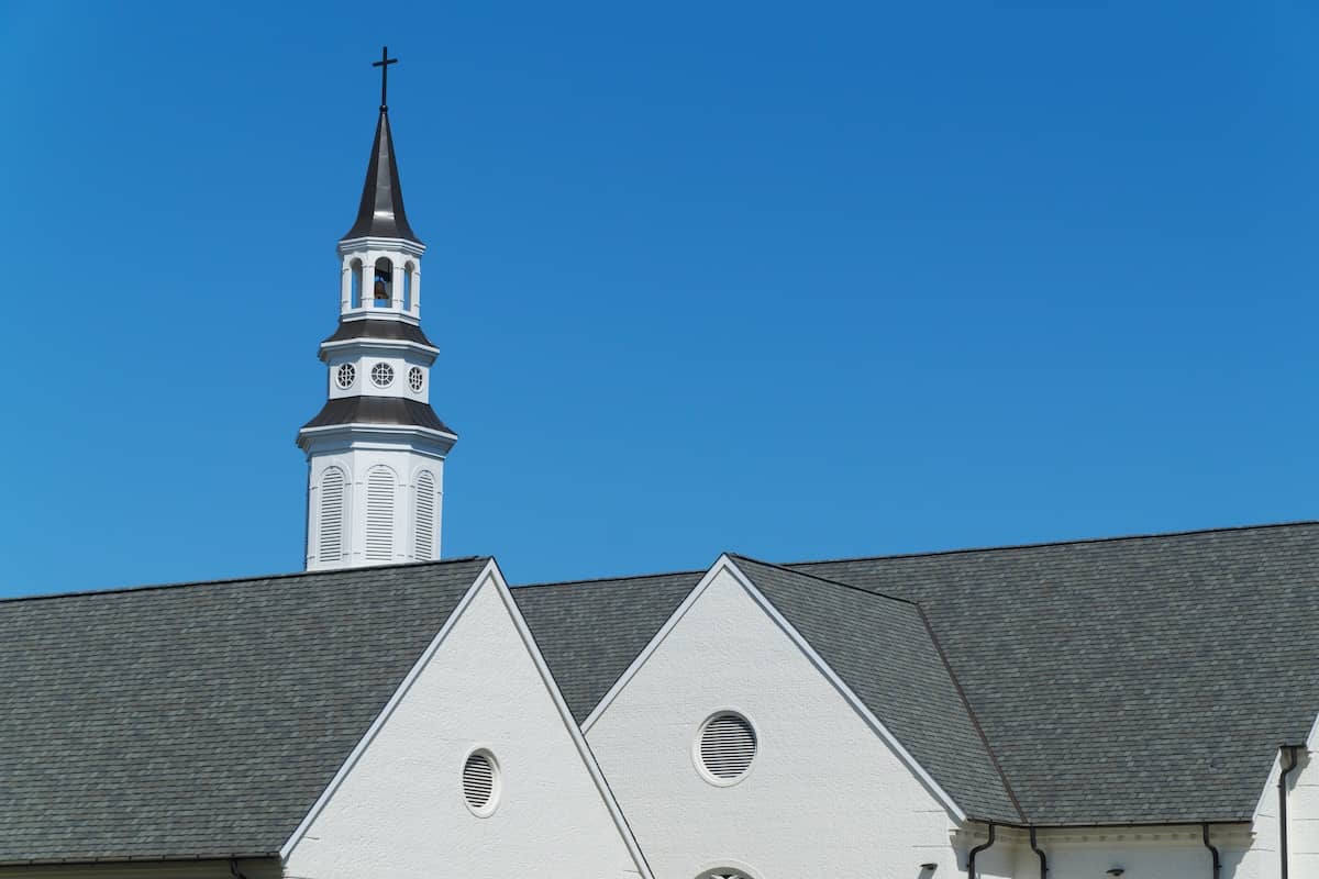 The image shows the roof and steeple of a white church against a clear blue sky. The steeple is topped with a cross and has a series of small windows. The building features triangular gables and circular vents.