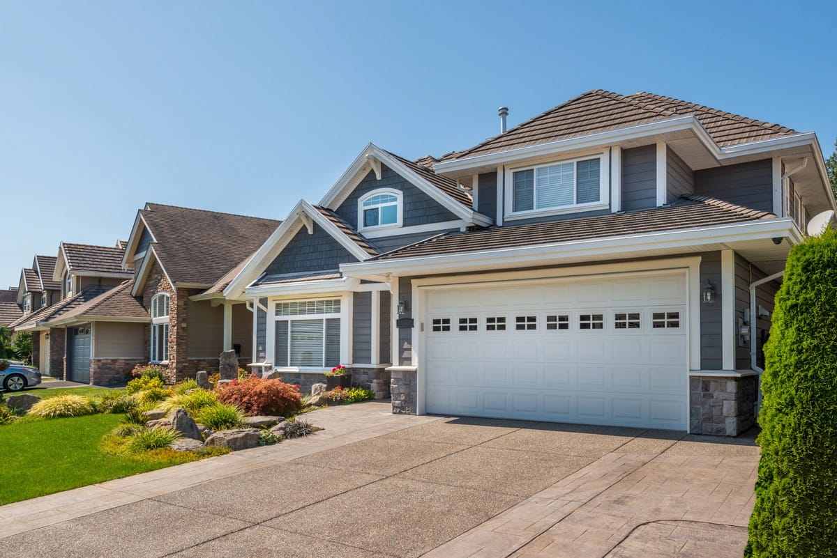A suburban street features a row of modern, two-story houses. The house in the foreground boasts a gray exterior with wooden shingles and a white garage door, complementing its well-maintained garden. Under the sunny skies, you might wonder, "How old is my roof compared to these?.