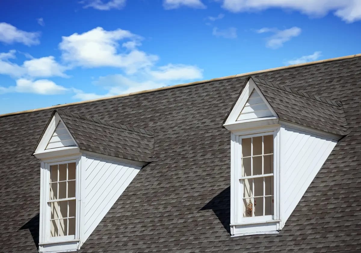 A roof with gray shingles features two dormer windows with white trim. Each window has multiple panes and the sky is bright blue with white clouds in the background.