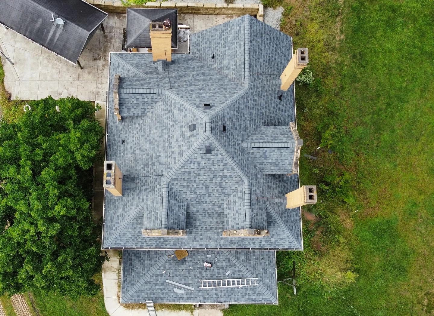 Aerial view of a large house with a complex, gray shingle roof. The roof has multiple chimneys and a ladder on it. Surrounding the house are green grassy areas and trees. A smaller structure is partially visible nearby.