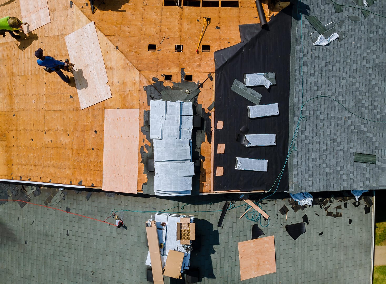 Aerial view of a roof under renovation. Two workers are placing plywood and tar paper, with tools and materials scattered around. Shingles are partially installed on the right side. A ladder leans against the roof.