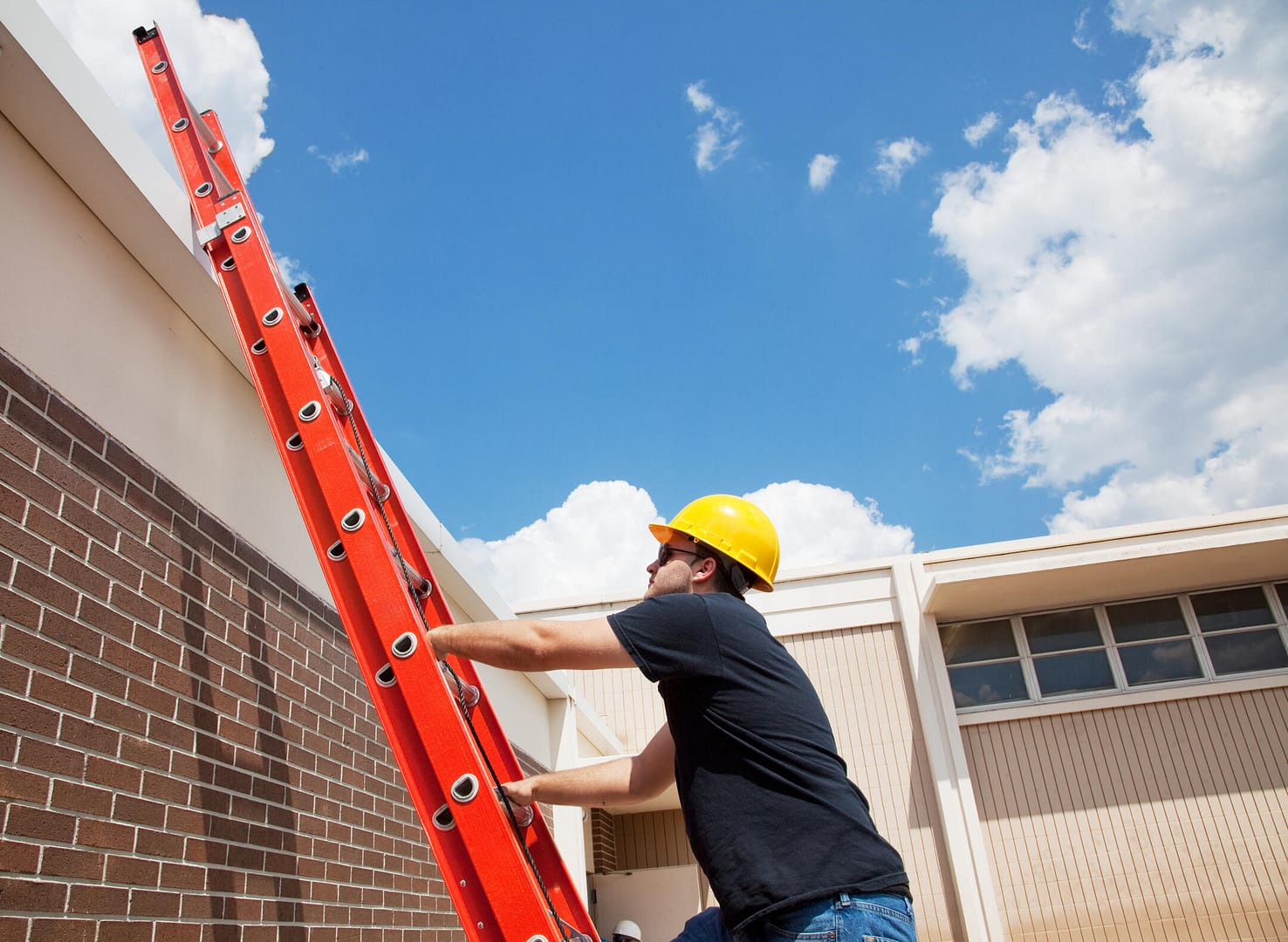 A worker in a yellow hard hat and black shirt climbs a red extension ladder leaning against a brick building. The sky is clear with a few clouds.