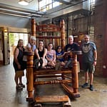 A group of six people smiling and posing on and around a giant wooden rocking chair indoors. The setting has a casual and industrial feel with visible brick walls and metal beams.
