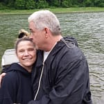 A man in a black jacket is embracing a woman in a black hoodie next to a river. The woman is smiling, and behind them is the water and a forested area. The scene appears to be outdoors and peaceful.