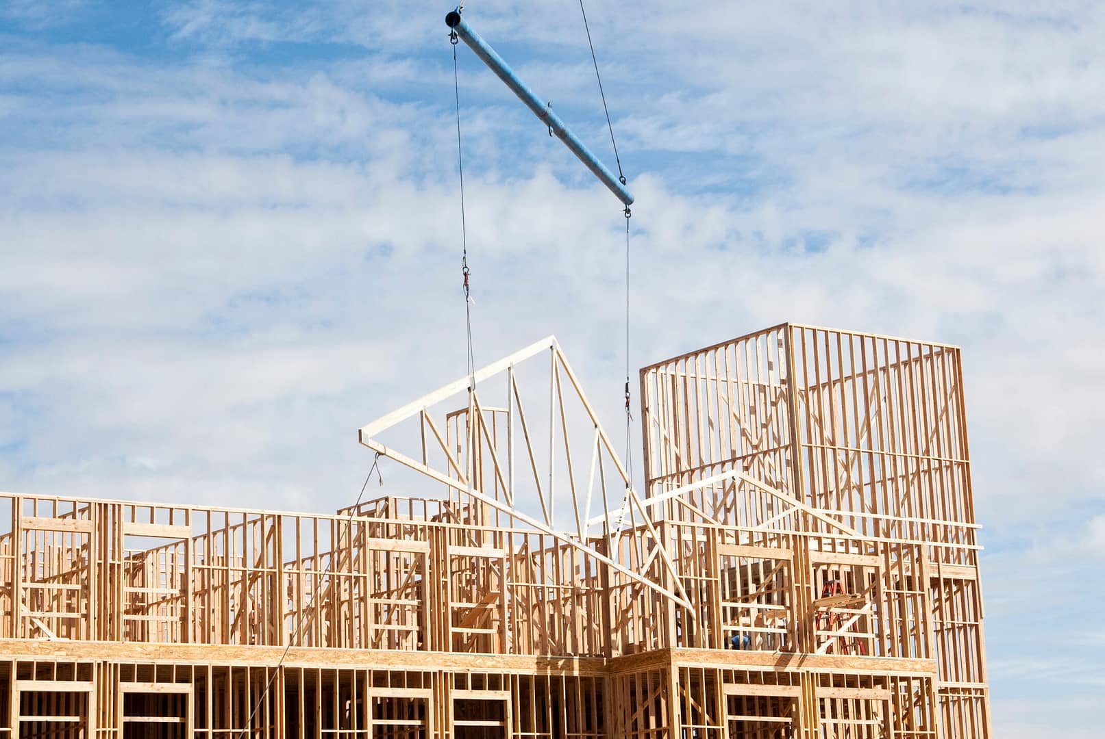 A wooden building under construction with a crane lifting a triangular roof truss into place. The frame consists of wooden beams, set against a partly cloudy blue sky.