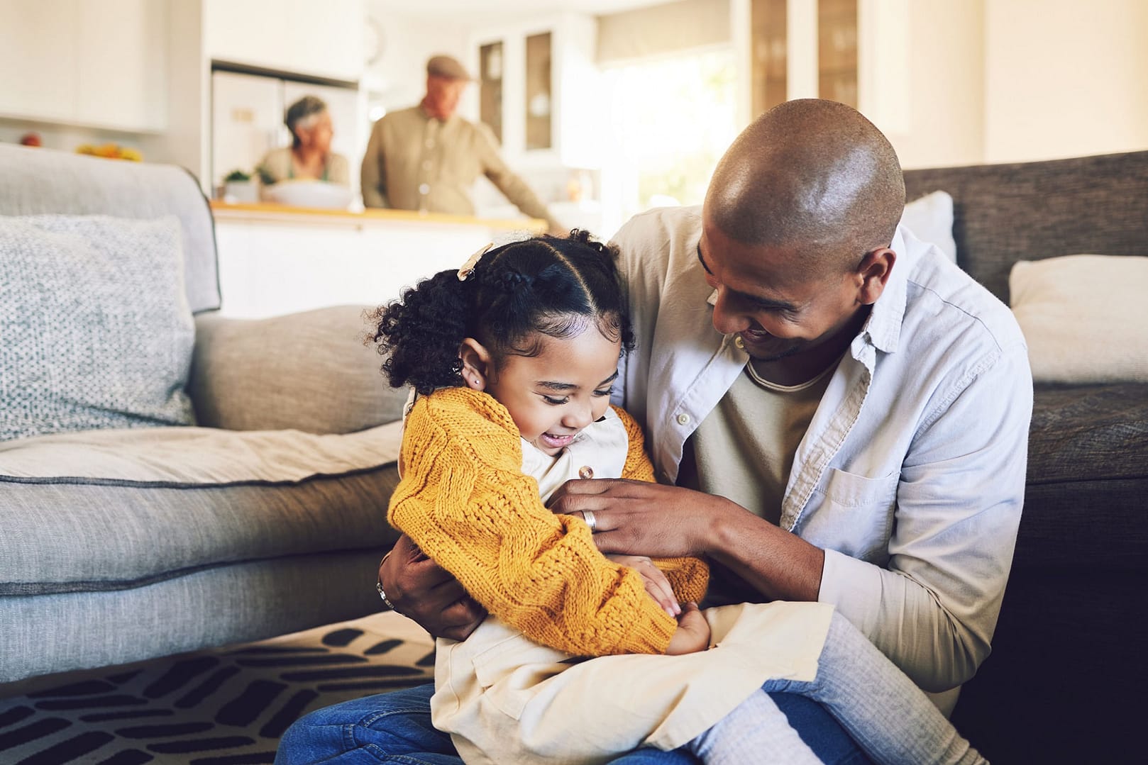 A father and daughter laugh together while sitting on the floor in a cozy living room. The young girl wears a yellow sweater. In the background, an older couple stands in the kitchen, smiling.