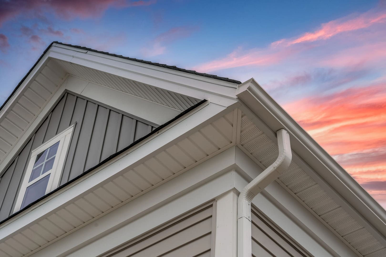 Close-up of a modern house's rooftop with white siding and a visible gutter against a colorful sunset sky featuring pink and orange clouds.