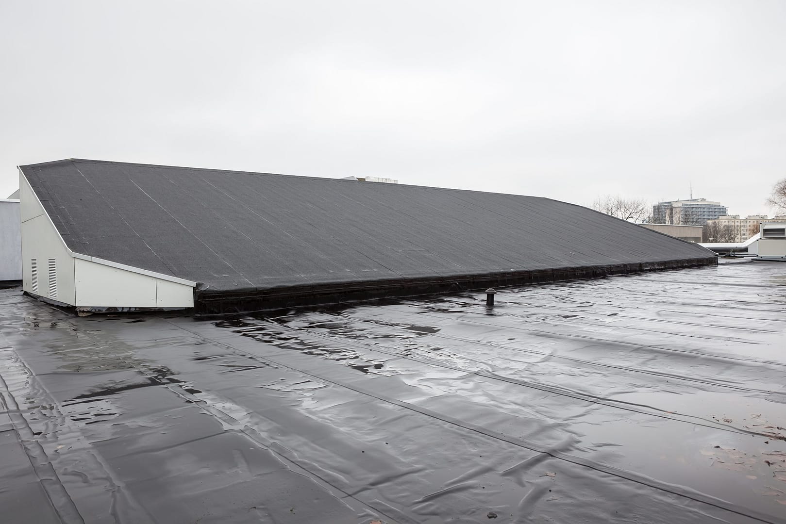 Flat rooftop covered with black waterproof material. A large, pitched structure is centered on the roof. The sky is overcast, and a few distant buildings are visible on the horizon.
