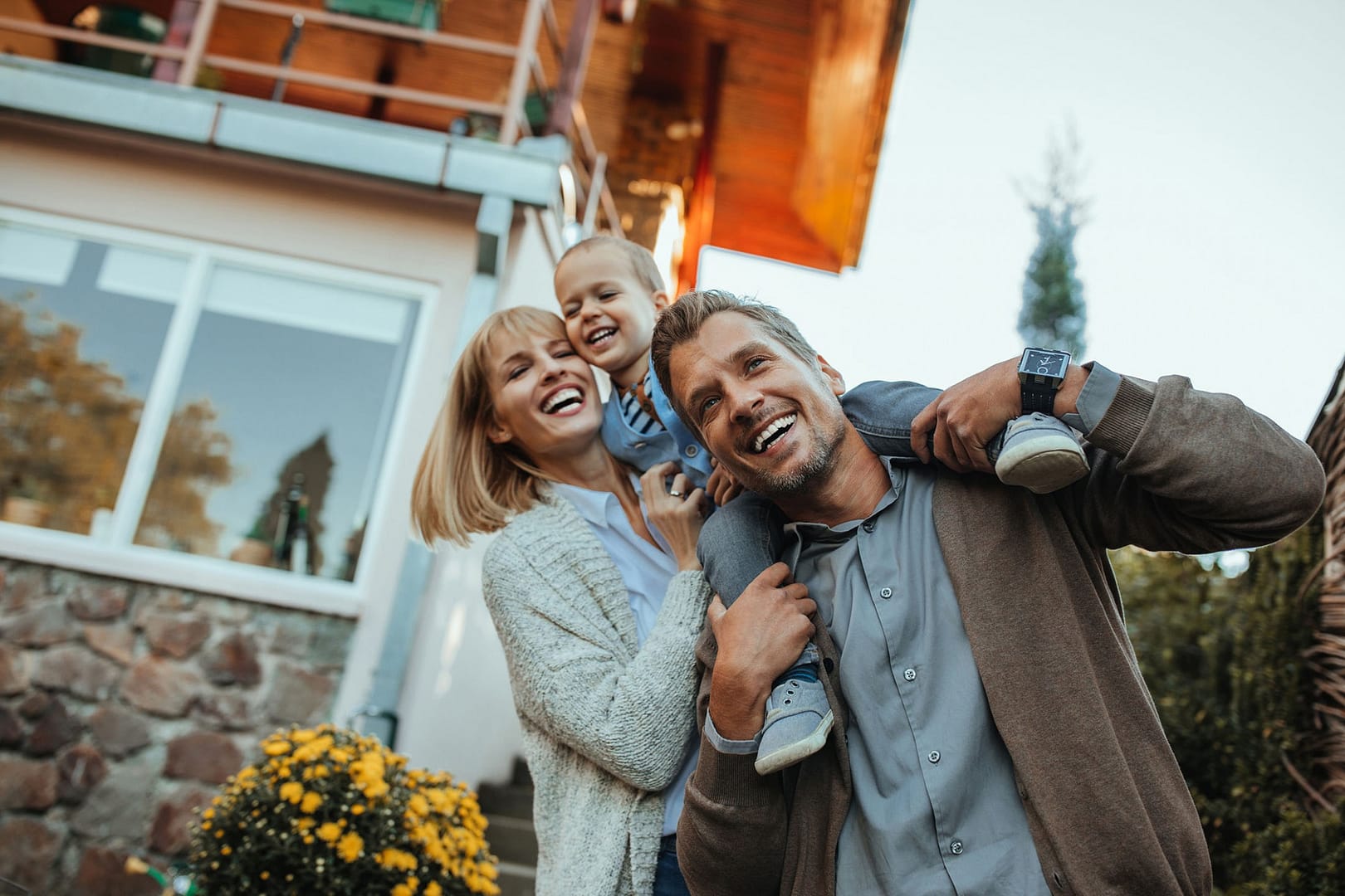 A happy family of three stands outside their home. The man carries the child on his shoulders, while the woman stands close, smiling. They are casually dressed, and there are yellow flowers and a stone wall in the background.