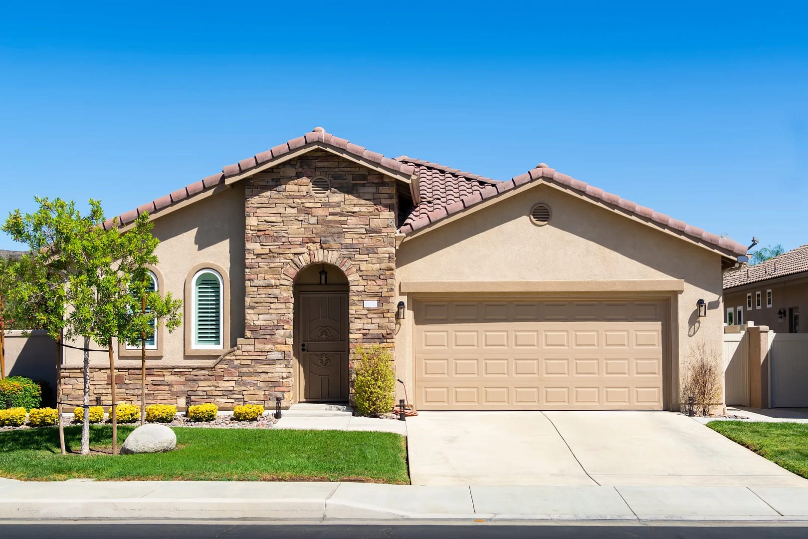 A single-story suburban house with a stone facade, arched entrance, and a two-car garage showcases stunning house and roof color combinations. The front yard features a trimmed lawn, small tree, and shrubs under a clear blue sky.