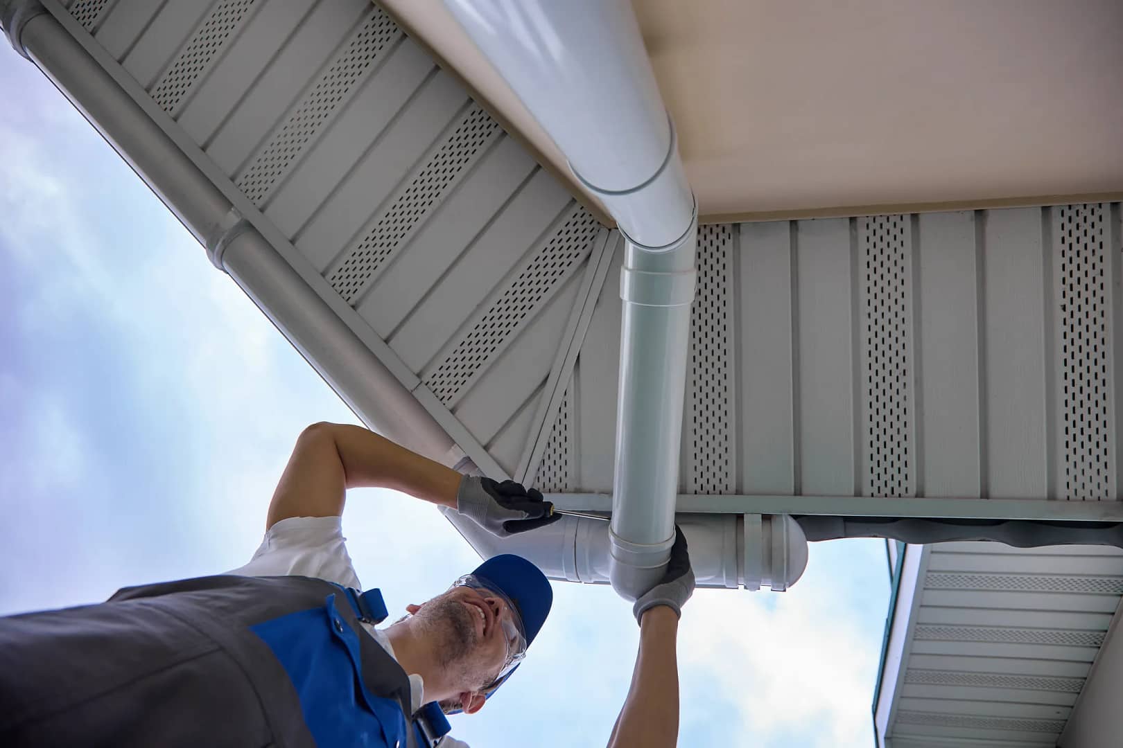 A worker in a blue cap and gloves is diligently repairing a white rain gutter under the eaves of a house, potentially saving on gutter replacement costs. Captured from a low angle, the image highlights the roof and sky in the background.