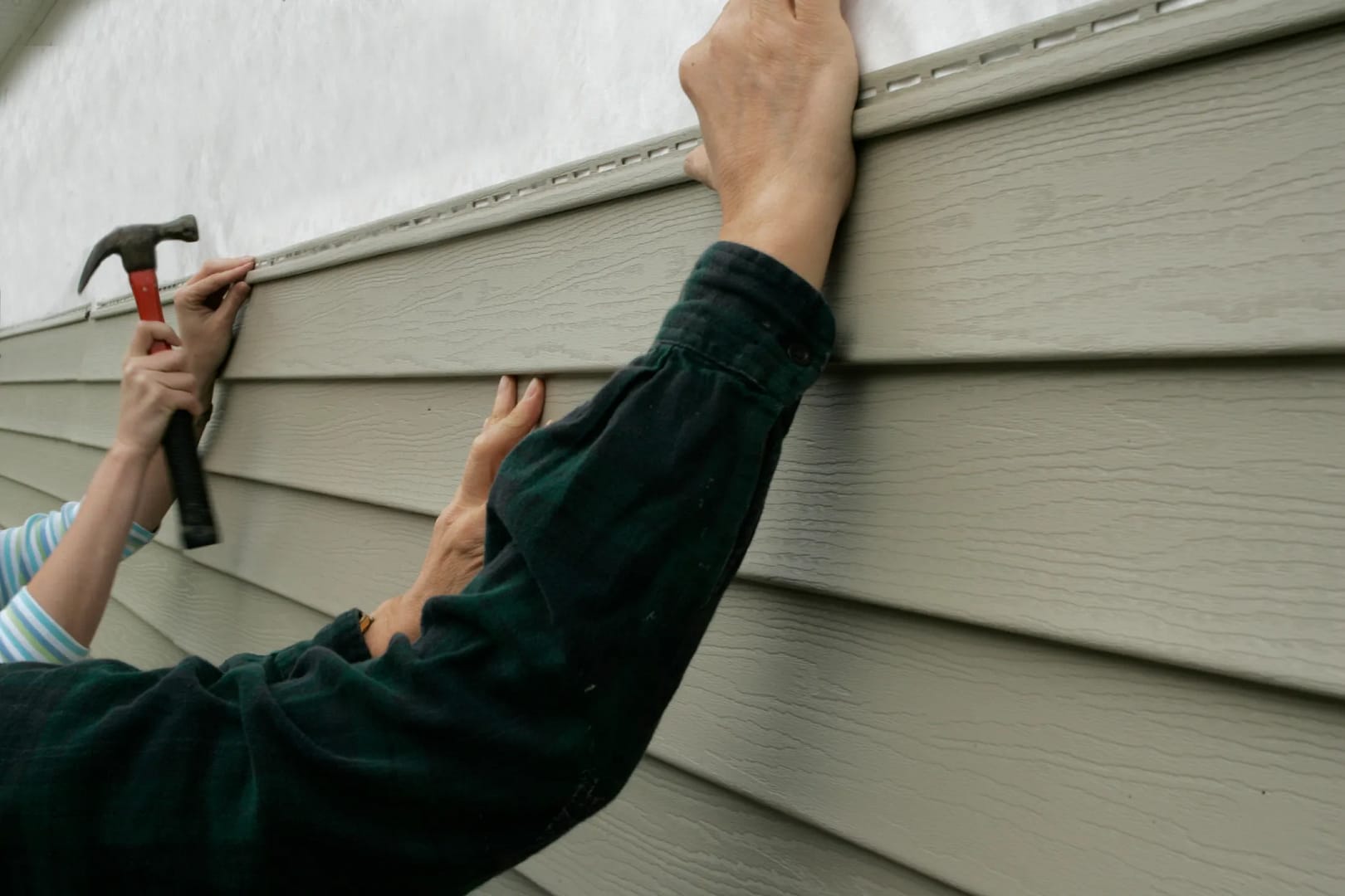 A person wearing a dark green plaid shirt installs light gray James Hardie siding on a house wall, securing it with nails using a hammer. Another individual in blue stripes offers assistance in the background.