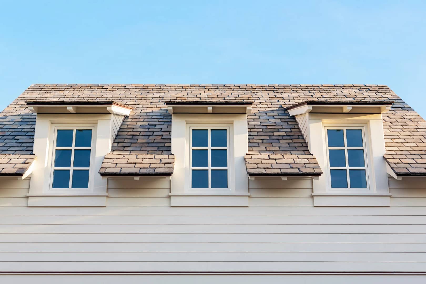 A house roof with three dormer windows featuring white frames and shutters. The exterior is clad in light-colored siding, hinting at recent siding replacement cost investments, and the roof is covered with brown shingles. The sky is clear and blue.
