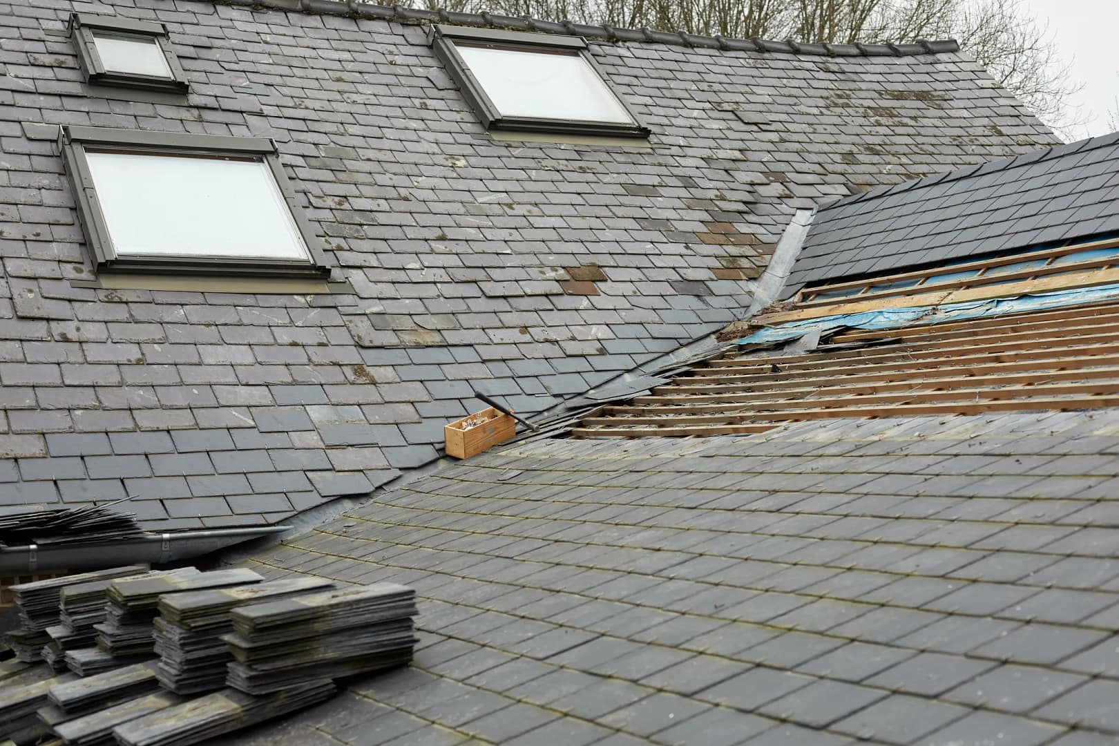 A roof with gray slate tiles, partially removed to reveal wooden beams, prompts the question: how long does a roof last? Two skylight windows peek through. A stack of tiles rests on the left, while blue tarp covers gaps. Trees serve as a natural backdrop.