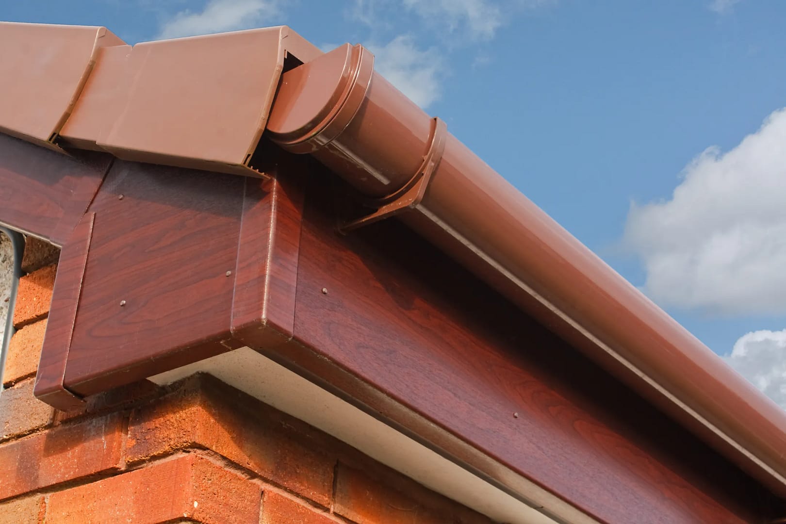 Close-up of a brown rain gutter and downspout attached to the eaves of a brick house, with a blue sky and clouds in the background. The wooden fascia board beneath the gutter matches the brown color of the system, showcasing recent fascia repair with seamless integration.