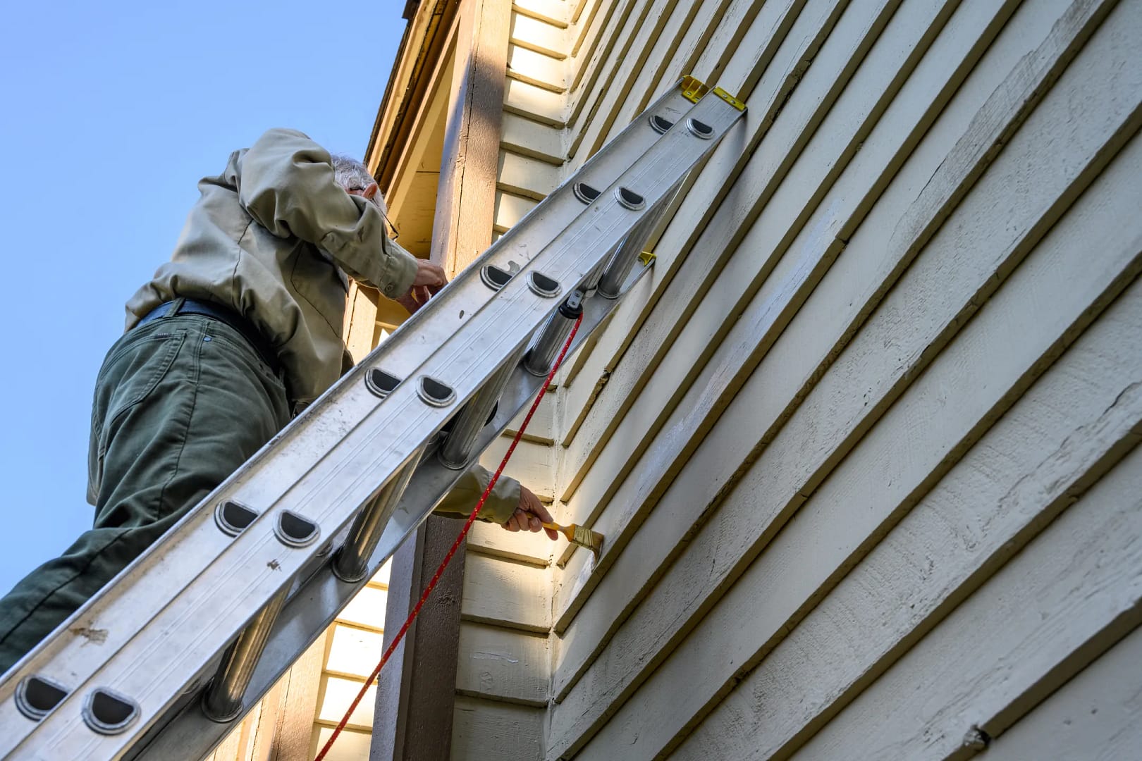 A person stands on a ladder, meticulously painting the exterior trim of a house adorned with beige vinyl siding. The sky is clear and blue above them as they work, wearing protective gloves and casual outdoor clothing.
