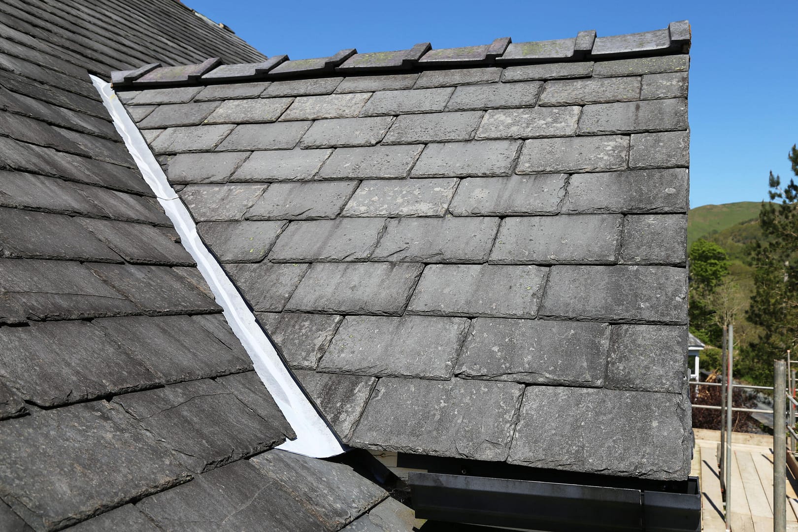 A close-up view of a slate roof with a valley, showing overlapping dark gray tiles and a strip of metal flashing for waterproofing. Clear blue sky in the background.