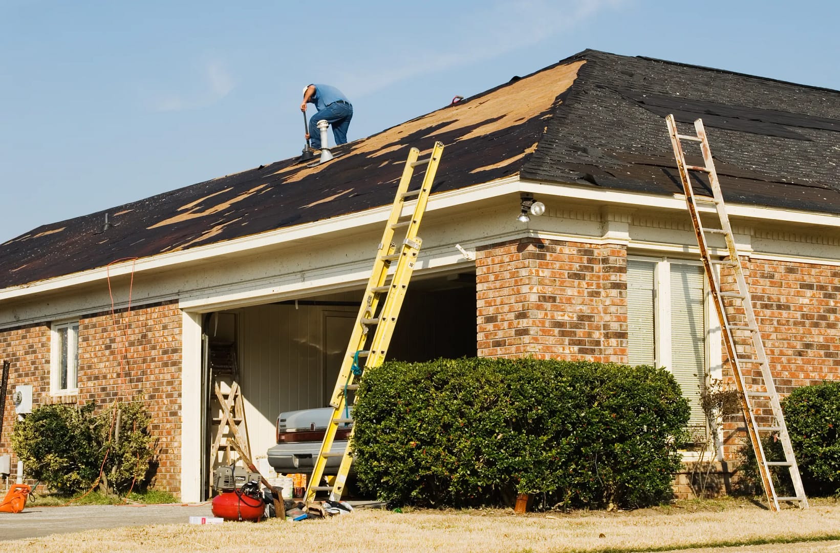 A person tackles a DIY roof repair on a brick house under renovation. Two ladders are propped against the roof, and the garage door is open, revealing tools and equipment inside. A blue sky serves as the backdrop for this hands-on project.
