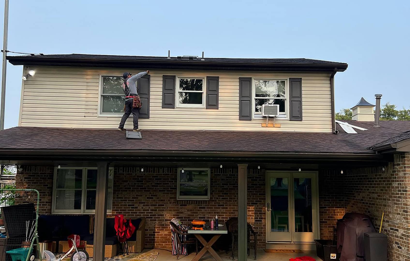 A person in work attire climbs a ladder to reach a second-story window of a brick house. The house has a covered patio with outdoor furniture and a grill. The sky is clear, suggesting a sunny day.