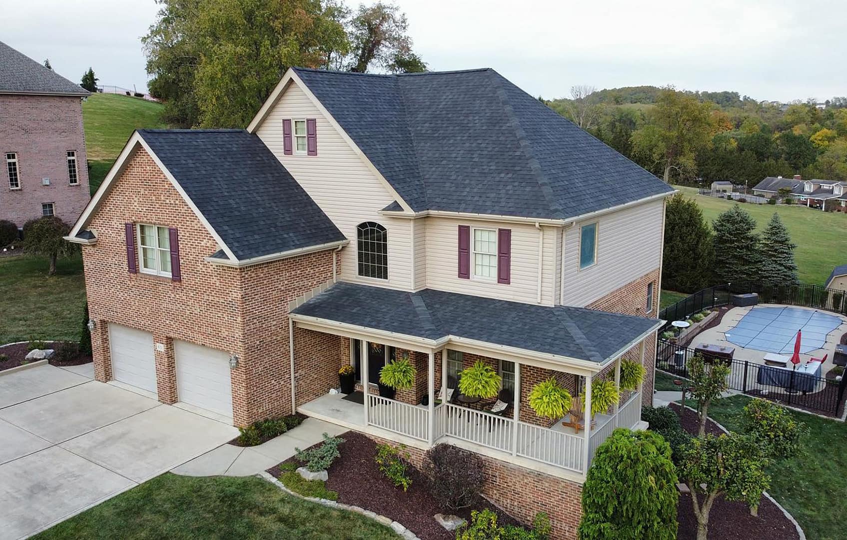 A two-story suburban house with a combination of brick and beige siding. It features a covered front porch with hanging plants, a two-car garage, and a neatly landscaped yard. A fenced pool is visible in the backyard.
