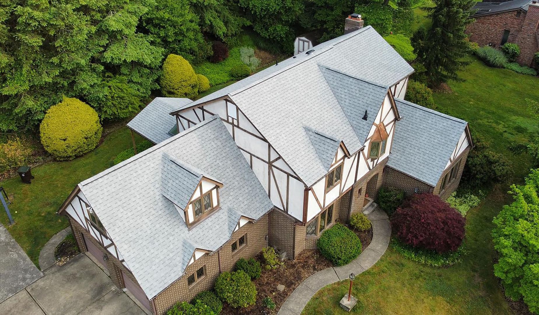 Aerial view of a Tudor-style house with a gray roof, brick walls, and decorative wooden beams. The property is surrounded by lush greenery and a landscaped garden with shrubs and trees. A paved driveway leads to the house.