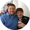 Two people sitting closely together in a bright setting, with one person holding a glass of dark beverage. Both are smiling, dressed casually, and appear to be enjoying a social moment.