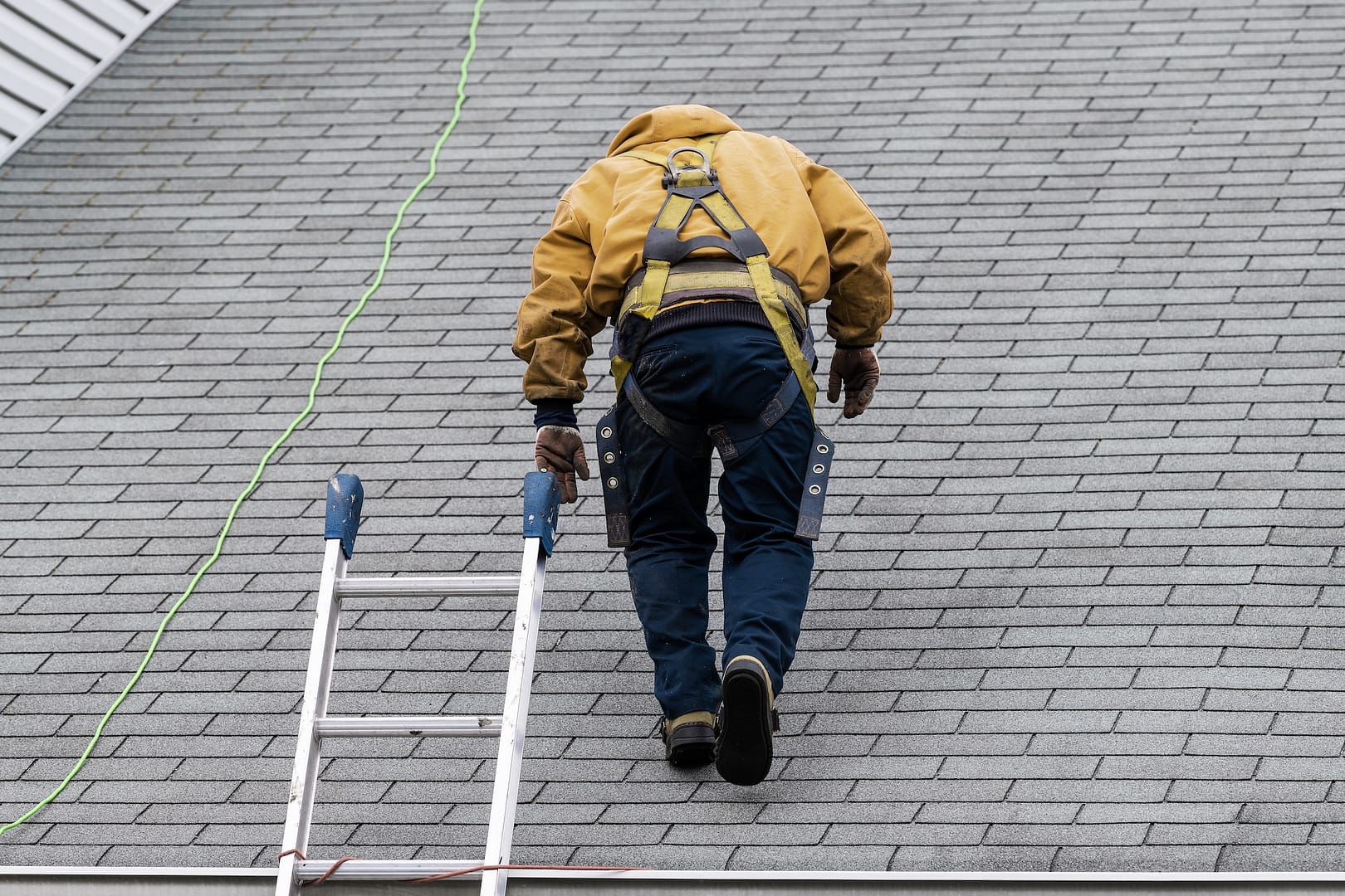 House during day with gray color Single Family Home and construction man in yellow uniform walking on roof shingles and ladder during repair
