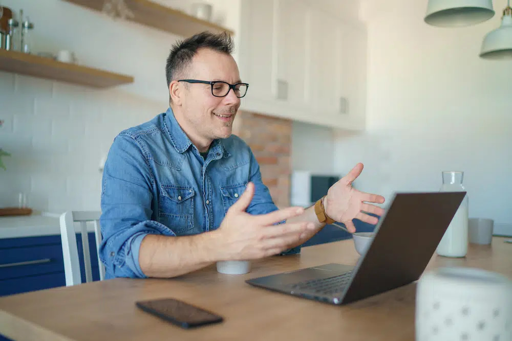 On the Hook Agency website, a man in a denim shirt is actively engaged in a video call discussing Phoenix web design. His animated gestures complement his conversation. A smartphone sits on the table, while kitchen shelves and a brick accent wall provide an inviting backdrop.
