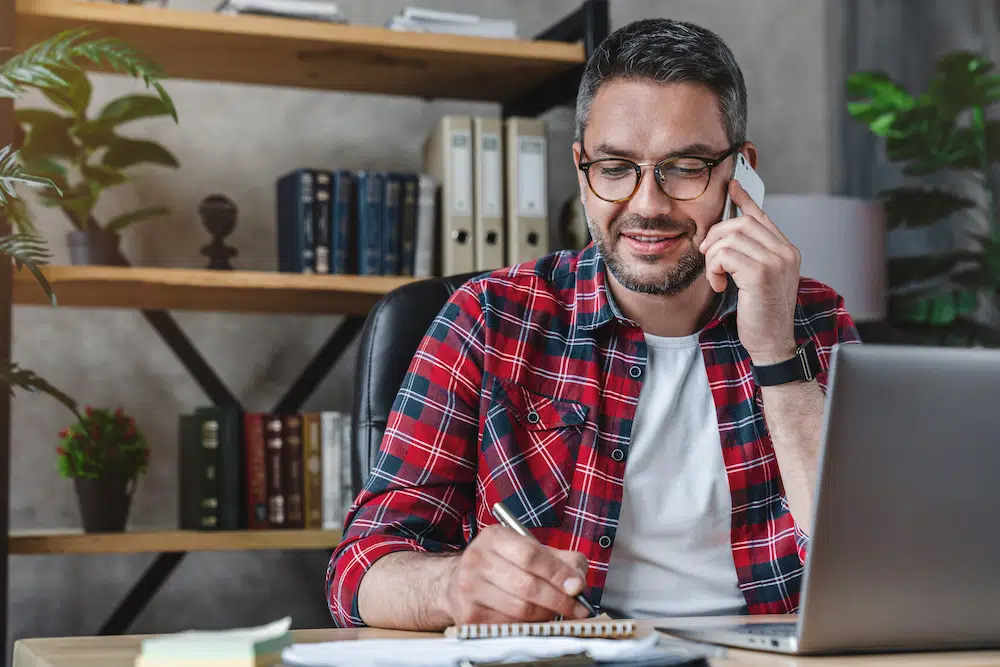 On Hook Agency's website, a man in glasses and a red plaid shirt is intently engaged in a conversation about Saint Louis SEO on his smartphone. As he takes notes in his notebook, an open laptop rests on his desk. The scene is complemented by shelves adorned with books and plants, adding an inviting touch to the setting.