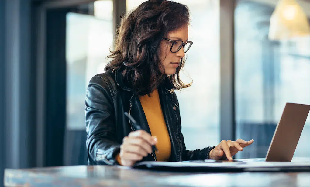 A focused individual with long hair and glasses, dressed in a black leather jacket and mustard yellow top, diligently works on Los Angeles web design projects at a table. With pen in hand, they engage deeply with their laptop against a backdrop of large, blurred windows.