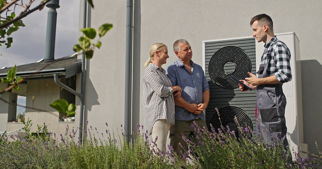 A technician demonstrates an outdoor heat pump unit to a couple in a garden, amid vibrant plants and flowers. This setting, enhanced by the backdrop of a building with visible pipes and windows, offers an engaging live review experience for SearchLight Digital.