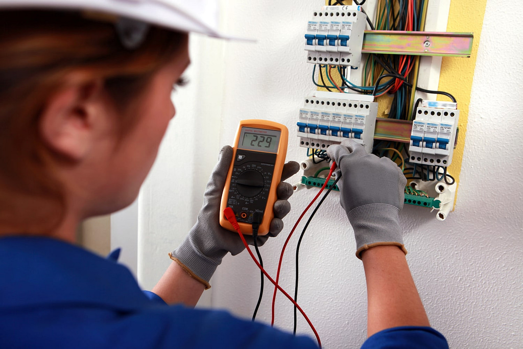 Electrical company names - electrician names - A technician in a hard hat and gloves uses a digital multimeter to measure 229 volts at an electrical panel with exposed wires and circuit breakers. This task underscores the expertise required in high-demand trades.