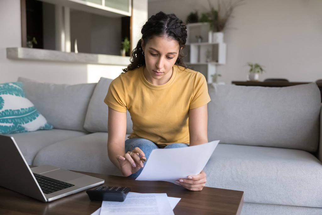 A woman in a yellow shirt reviews HVAC and plumbing financing documents on a couch. A laptop, calculator, and additional paperwork are organized on the wooden table before her. The bright, modern room features plants and a neutral color scheme.