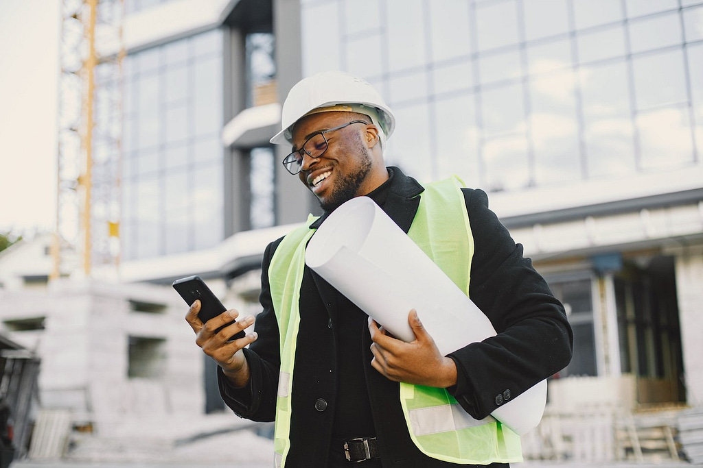 A construction worker, equipped with a hard hat and high-visibility vest, confidently stands before a modern building under construction. With blueprints in hand, he efficiently checks social media management tools on his smartphone.