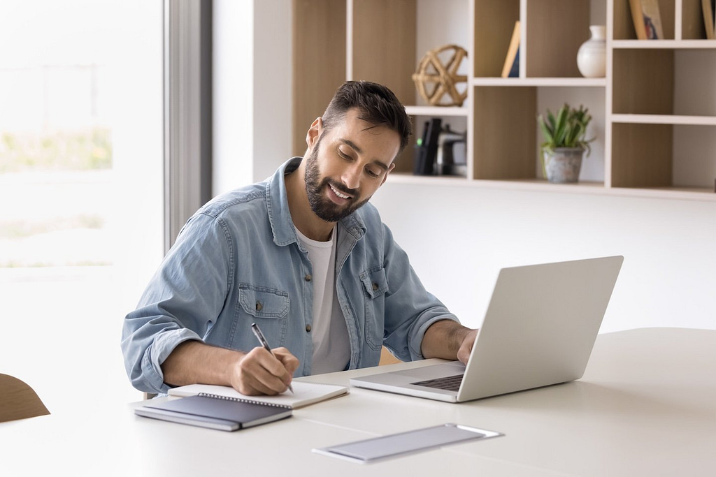 A bearded man smiles while diligently working on his laptop and taking notes from a practical guide. Surrounded by a well-lit room with decorative shelves and a plant, he focuses on calculating the value of a business in the heating and air conditioning industry.