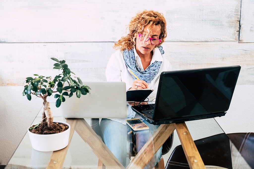 In a bright and modern indoor space, an individual with curly hair and distinctive pink glasses is focused on writing in a notebook, accompanied by two open laptops looking at hvac ecommerce and a potted plant.