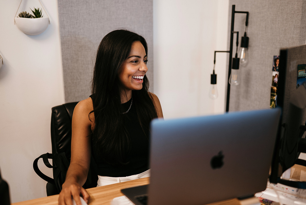 A woman with long dark hair in a black sleeveless top sits at a desk, deeply focused on managing Google Ads for home services. Her workspace is highlighted by gray acoustic panels, a modern lamp with exposed bulbs, and small potted plants.