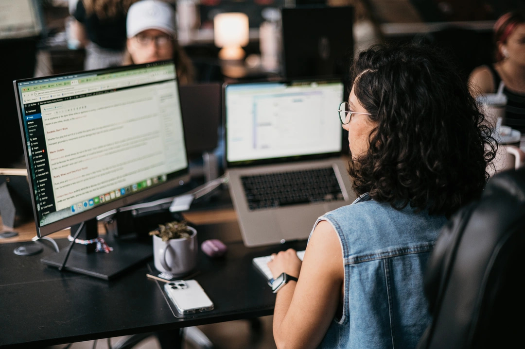 A woman with curly hair and glasses diligently works at her desk, efficiently managing Fire Restoration data on a large monitor and laptop. Her workspace is complemented by a smartphone and a potted plant, creating a lively atmosphere. A colleague is visible in the blurred background of this office setting.
