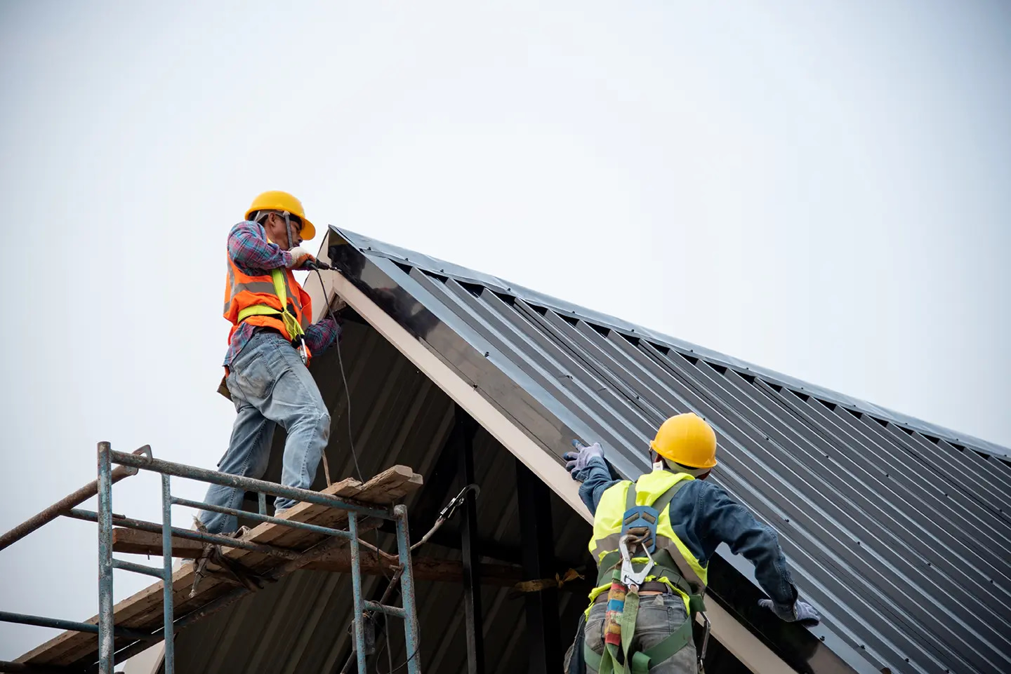 Two construction workers, equipped with safety gear and helmets, efficiently tackle a roofing project on a sloped metal surface. One worker secures materials from the scaffolding while the other directs operations from above. Set against a clear sky, this scene exemplifies their expertise in LSA services.