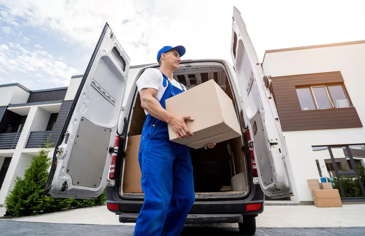 A delivery professional, clad in blue coveralls and a cap, efficiently unloads a company-branded cardboard box from an open van amidst a residential neighborhood. With other boxes visible inside the vehicle and under partly cloudy skies, the streamlined moving process is well underway.
