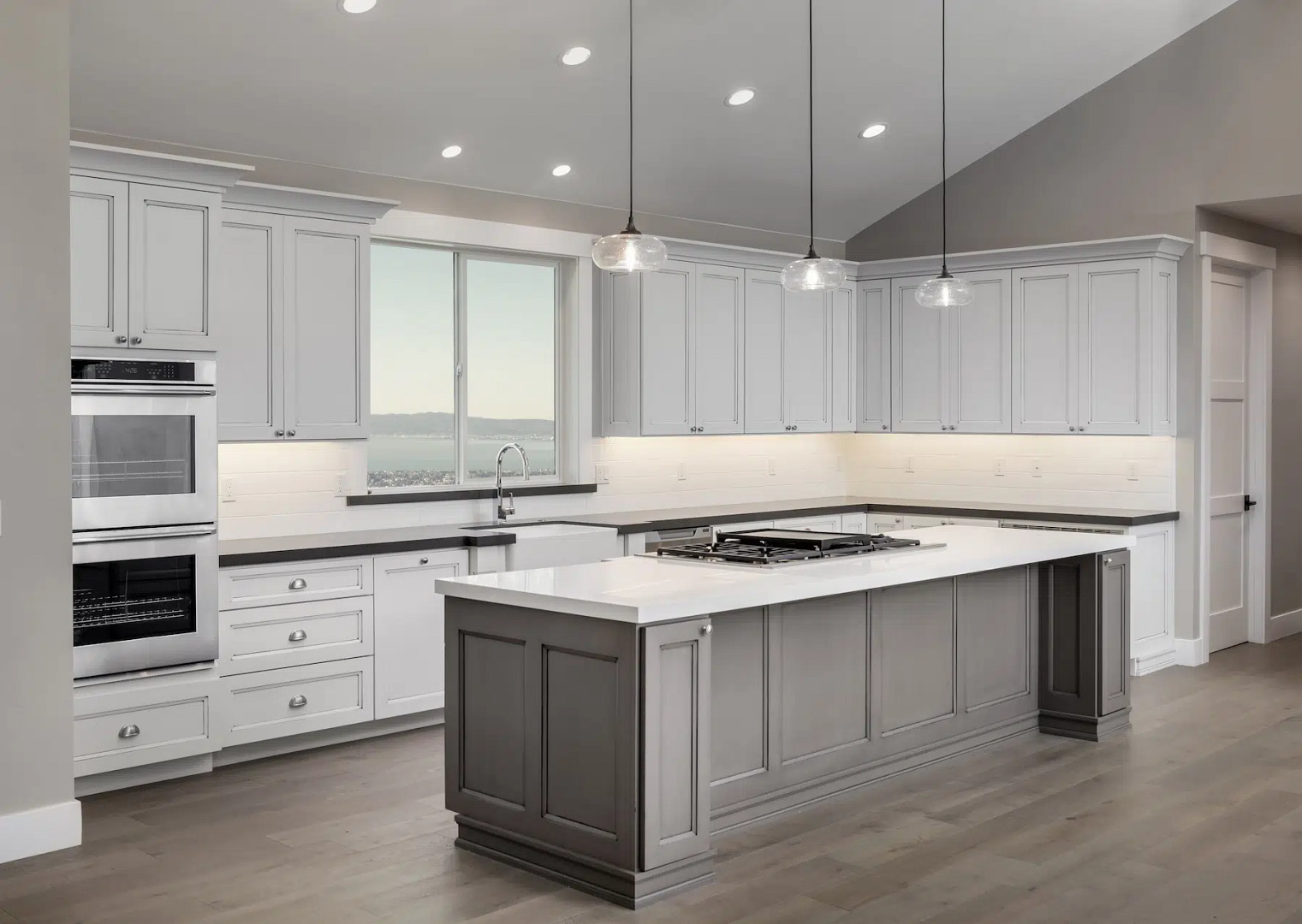 Explore our modern kitchen design featuring sleek white cabinets and a spacious gray island topped with a pristine white countertop. Seamlessly integrated stainless steel appliances, including a double oven, enhance functionality. Elegant pendant lights artfully illuminate the island, complemented by a window view that welcomes the outside world in.