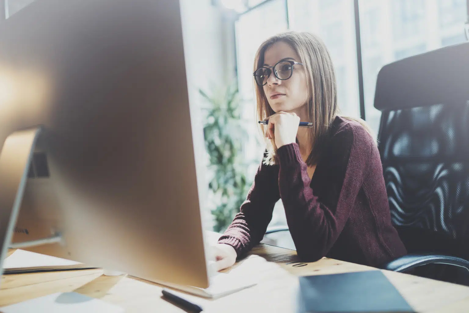 A focused woman with glasses sits at her desk, analyzing SEO strategies on her computer screen. She contemplates the data, chin in hand and pen poised for notes. Bathed in natural light, the room highlights her workspace where an open notebook labeled "Madison" is filled with insights and strategies.