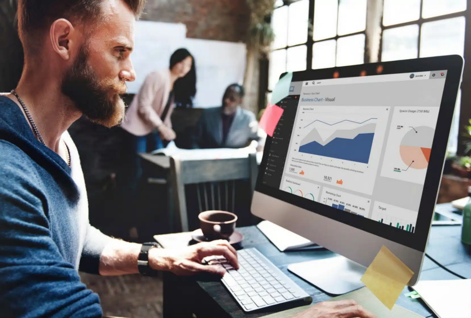 In a vibrant San Jose office, a man concentrates on SEO graphs and charts displayed on his computer. Nearby, two colleagues collaborate in the modern workspace, characterized by large windows and an array of papers scattered across the desk.