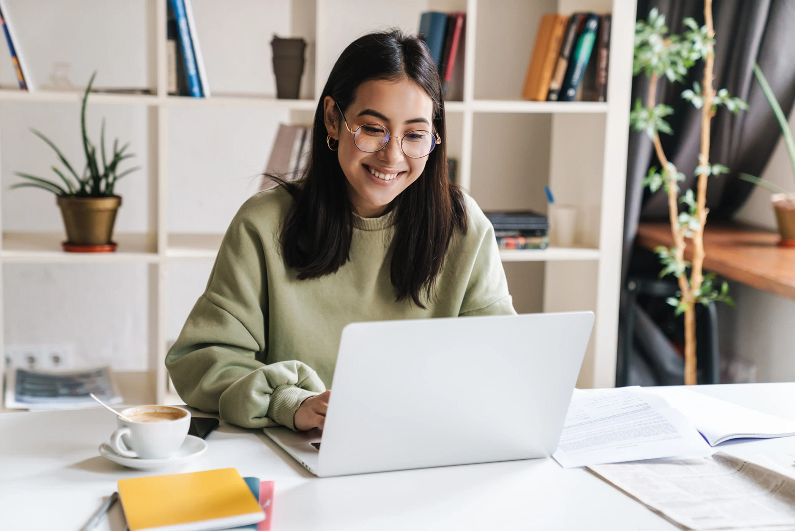 A woman in glasses and a green sweater smiles as she diligently works on a Houston web design project at her desk. Her workspace features books, papers, and a cup of coffee, while a plant-filled bookshelf adds to the cozy atmosphere.