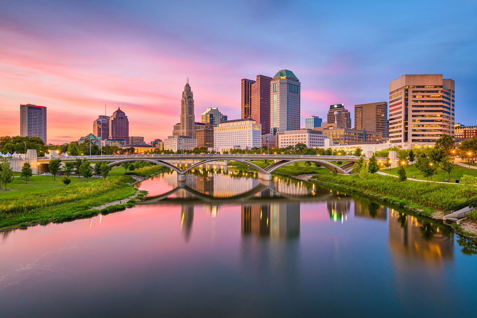 Experience the dynamic blend of modern and historic architecture in Columbus, Ohio. At sunset, the vibrant cityscape reflects pink and orange hues on the calm river, embodying a bustling hub for Columbus SEO that seamlessly bridges innovation with tradition. Discover how Hook Agency can elevate your digital presence in this thriving metropolis.